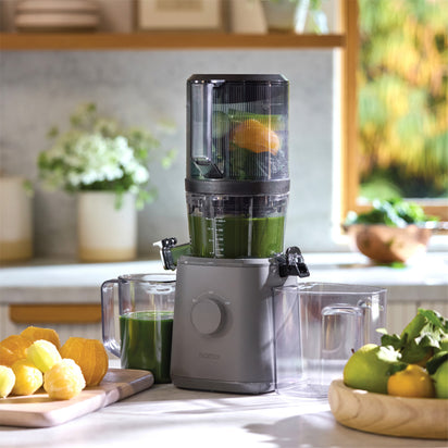 The Nama J2 cold press juicer, filled with leafy greens and citrus slices, sits on a kitchen counter surrounded by juice containers, whole lemons, limes, and oranges with sunlight streaming through the window in the background.