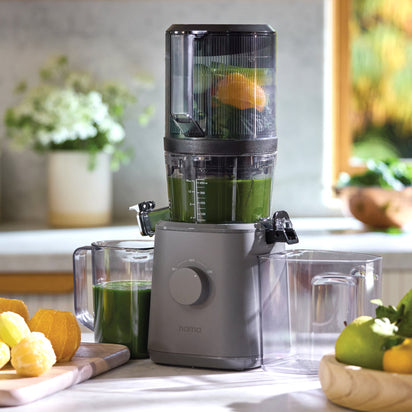 A Nama J2 juicer, filled with green juice, sits on the countertop between two clear containers. Lemons and leafy greens rest nearby, illuminated by natural light from a window.