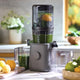 A Nama J2 juicer, filled with green juice, sits on the countertop between two clear containers. Lemons and leafy greens rest nearby, illuminated by natural light from a window.