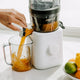 A hand operates the Nama J2 Plug Type I white cold press juicer, pouring fresh orange juice into a clear pitcher on a white countertop, with lemons and limes nearby.