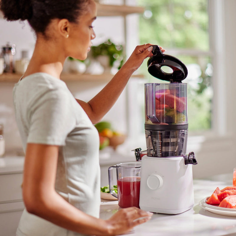 A woman places fruit into her easy-to-clean Namawell Nama J2 juicer in a bright kitchen. A glass of red juice sits on the counter nearby, and fresh produce is visible in the background.