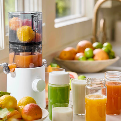 The Namawell Nama J3 cold press juicer sits on a kitchen counter, filled with oranges and grapefruits. Around it are jars and glasses of fresh juice, with bowls of assorted citrus fruits near a sunlit window in the background.