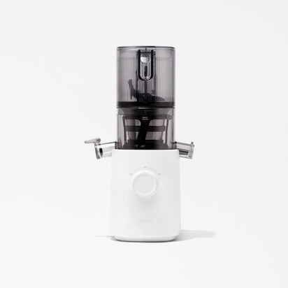The Nama J3 Plug Type I is a sleek, modern cold press juicer with a transparent upper chamber and a white base with a central control knob, shown against a plain white background.
