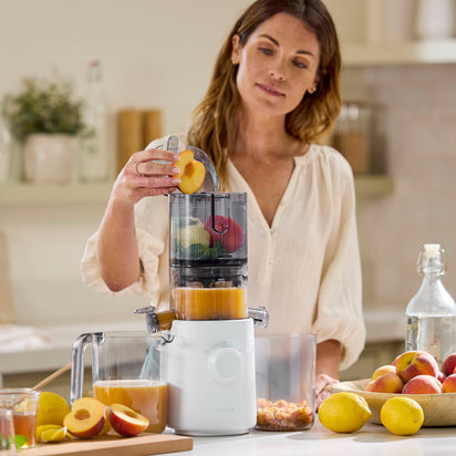 A woman in a light blouse uses the Nama J3 cold press juicer on her kitchen counter, placing fresh peaches inside while jars of homemade peach juice and whole peaches surround her.
