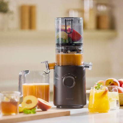 The black Nama J3 cold press juicer pours fresh juice from sliced fruits into a pitcher, while glasses with ice and garnishes sit nearby alongside peaches and fruit slices on a cutting board in a sunny kitchen.