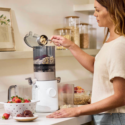 A woman in a kitchen adds cashews from a measuring cup to a vertical white food processor. Fresh strawberries and dates sit nearby, next to the easy-to-clean Nama J3 juicer and jars of pantry items in the background.