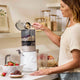 A woman pours cashews from a measuring cup into the Nama J3 Plug Type G juicer on a modern counter, with strawberries and jars of grains nearby, showcasing the easy-to-clean appliance in her sleek kitchen.