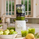 A white Nama J3 Plug Type G cold press juicer sits on a kitchen counter, processing green guavas. Fresh juice collects in a glass and pitcher, with guavas, lemons, and juice nearby. A window appears in the background.