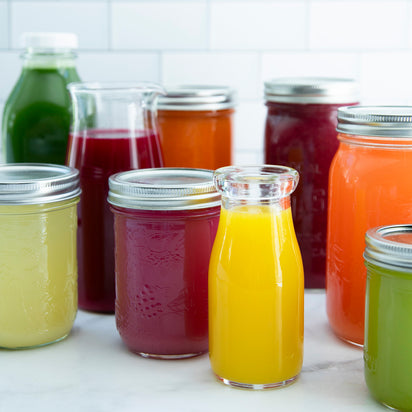 An array of vibrant juices in clear bottles is displayed on a white surface with a tiled backdrop—great inspiration for Juicing for Beginners or anyone exploring plant-based recipes.