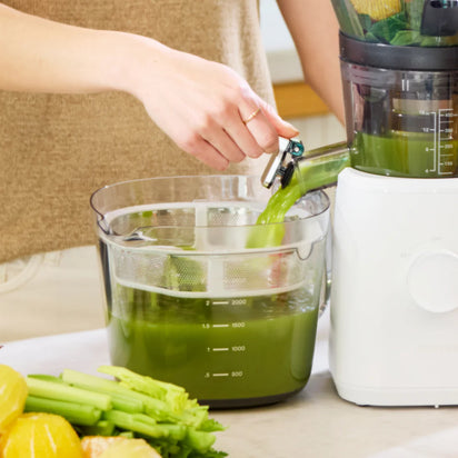 A person uses a white juicer to pour fresh green juice into a container, with celery, lemons, and greens on the counter. The Large Pitcher Set is nearby, providing convenient storage for juice made from healthy ingredients.