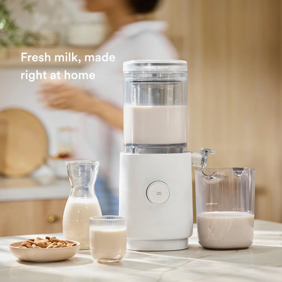 The Nama M1 by Nama is a countertop plant-based milk maker. Enjoy fresh nut milk in a pitcher, glass, and carafe with almonds nearby. A blurred person stands in the background. Text reads: Fresh milk, made right at home.