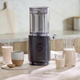 A sleek black Nama M1 sits on a kitchen counter amid glasses, a carafe of barista-quality milk, a ceramic cup, and grains. The bright, modern setting highlights this plant-based milk maker's stylish design.