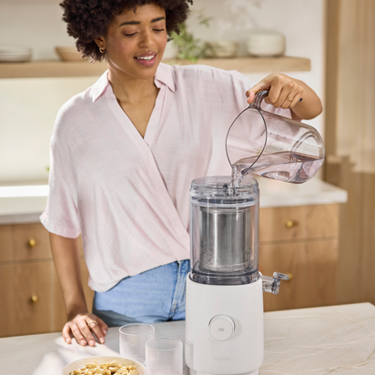 A woman in a light pink shirt pours water into a Nama M1 Plug Type E plant-based milk maker, with two empty glasses and a bowl of cashews nearby in a bright, modern kitchen ready for homemade nut milk.