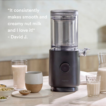 The Nama M1 by Nama sits on a kitchen counter with glasses of nut milk, bowls of nuts, and a mug. Text: "It consistently makes smooth, barista-quality milk and I love it!" – David J.