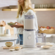 On a kitchen counter, the Nama M1 Plug Type G plant-based milk maker sits among bowls of nuts, oats, a glass bottle, and a clear pitcher. A woman holds a bowl in the softly lit modern kitchen—ideal for making fresh nut milk at home.