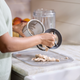 A person pours the contents from a Nama M1 plant-based milk maker onto a cutting board on a kitchen counter, with fruit in the background.