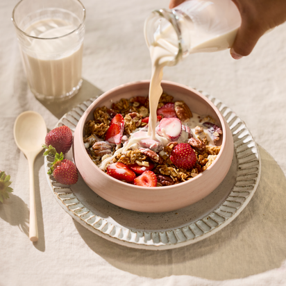 A hand pours nut milk made with the Nama M1 Plug Type I into a bowl of granola with strawberries and pecans. Beside the bowl are a glass of creamy milk, a wooden spoon, and two strawberries on a light tablecloth.