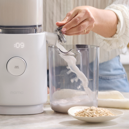 Using the Nama M1, a person pours fresh homemade nut milk from the blender container into a clear pitcher, with a bowl of dry oats nearby on the counter.