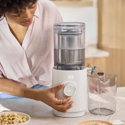 Wearing a light pink shirt, a person adjusts the dial on the Nama M1, a modern white plant-based milk maker with a digital display and transparent container, on a kitchen counter beside a clear pitcher and bowl of cereal.
