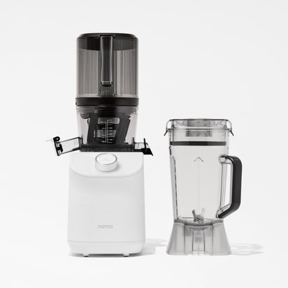 A white Nama C2 cold press juicer with a transparent top section stands beside a clear blender pitcher with a black handle, both set against a plain white background.