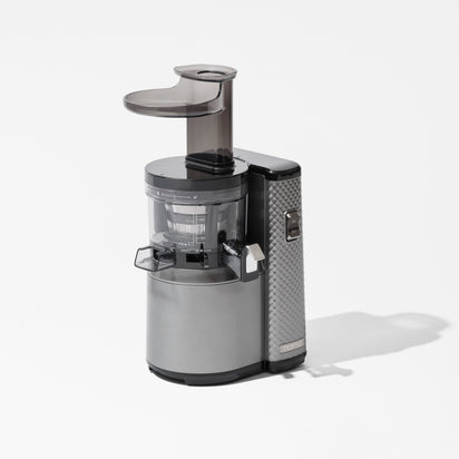 The Nama J1 is a modern cold press juicer in silver and black, featuring a transparent chamber and dark top chute, designed for easy cleaning and making nutrient-rich juices. Shown against a plain white background.