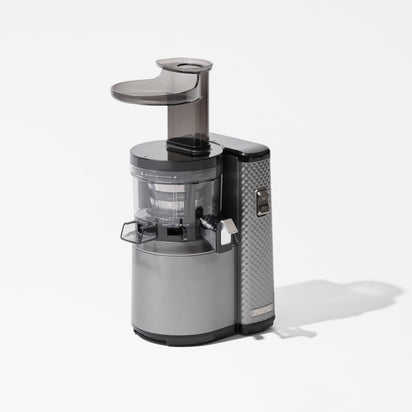 The Nama J1 Plug Type G is a sleek, modern cold press juicer featuring a transparent top chute, cylindrical silver body with textured accents. Easy to clean and ideal for nutrient-rich juices, it's shown against a plain white background.