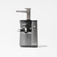 The Nama J1 Plug Type G is a modern silver and gray cold press juicer with a transparent container and textured panel, designed for easy cleaning and nutrient-rich juices. It stands upright on a white background.