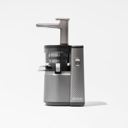 The Nama J1 Plug Type E is a modern vertical juicer in silver and black, featuring a transparent juice container and textured panel, displayed on a plain white background.