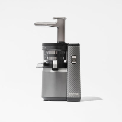 The Nama J1 Plug Type I is a sleek gray and black cold press juicer with a transparent chamber and textured side panel, designed for easy cleaning, shown against a plain white background.