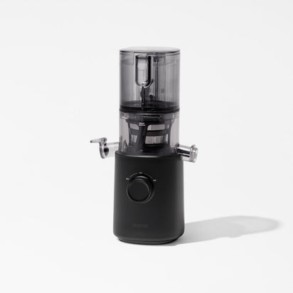 The Nama J3 Plug Type I is a modern black cold press juicer with a transparent top chamber and a single large dial on its base, pictured standing on a white surface with minimal shadow.