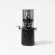 The Nama J3 Plug Type I is a modern black cold press juicer with a transparent top chamber and a single large dial on its base, pictured standing on a white surface with minimal shadow.