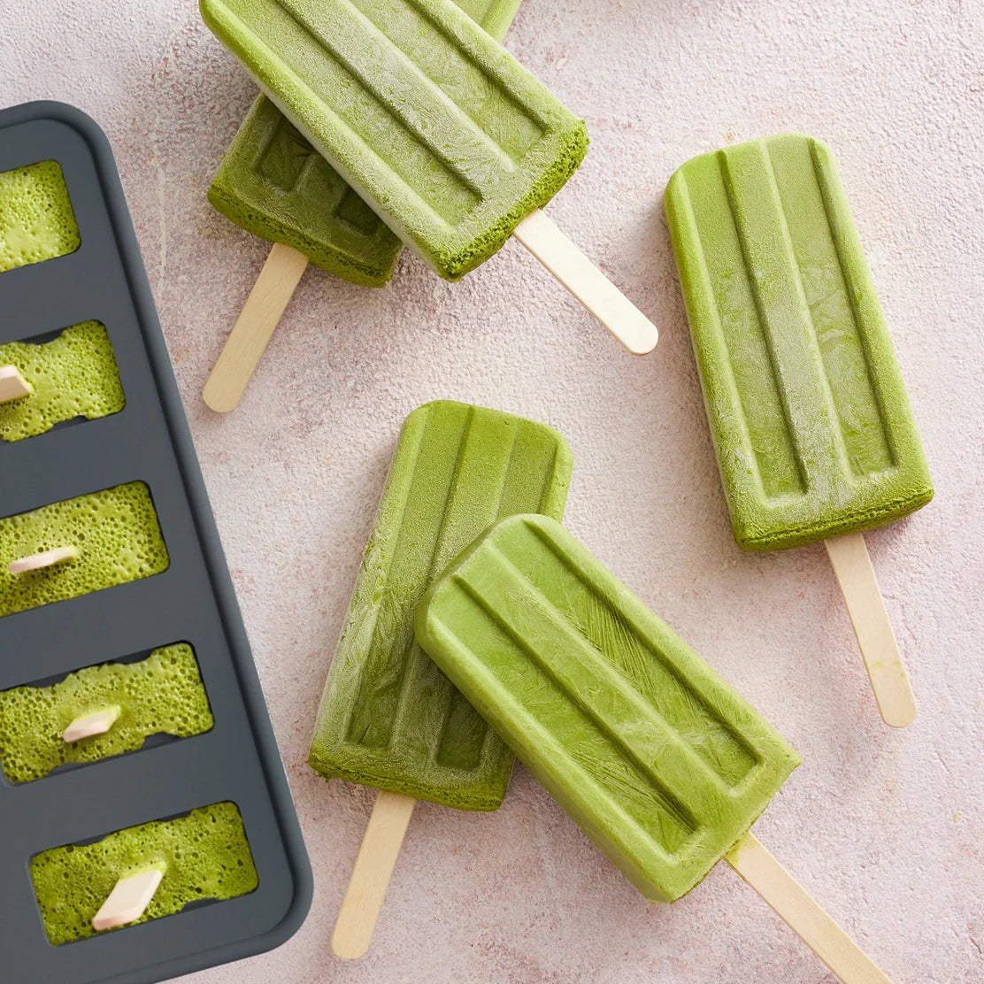 Four green popsicles made with the Nama Popsicle Mold rest on a light textured surface beside the black Nama mold, which is filled with more green popsicle mixture.