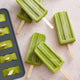 Four green popsicles made with the Nama Popsicle Mold rest on a light textured surface beside the black Nama mold, which is filled with more green popsicle mixture.