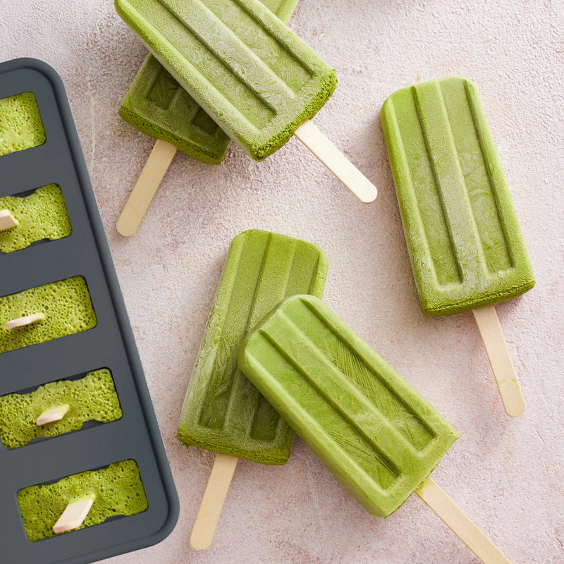 Four green popsicles made with the Nama Popsicle Mold rest on a light textured surface beside the black Nama mold, which is filled with more green popsicle mixture.