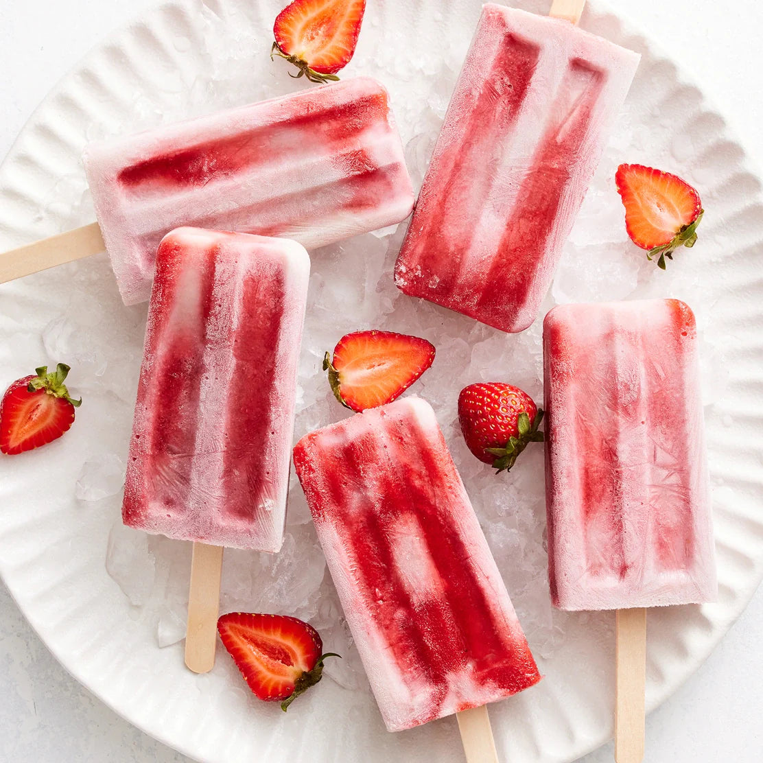 Five red and white striped popsicles made with the Nama Popsicle Mold are arranged on a plate of ice, surrounded by fresh halved and whole strawberries.