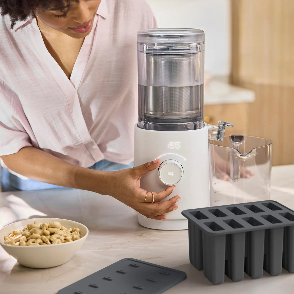A person adjusts a Nama Popsicle Mold on a countertop beside a bowl of cashews and two black silicone ice cube trays, suggesting food preparation in a bright kitchen.