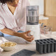A person adjusts a Nama Popsicle Mold on a countertop beside a bowl of cashews and two black silicone ice cube trays, suggesting food preparation in a bright kitchen.