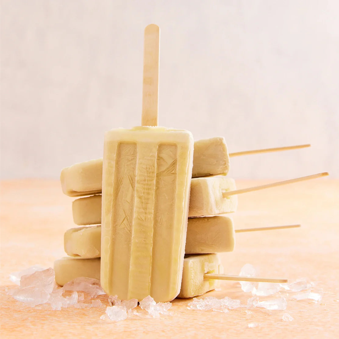 Six popsicles made with the Nama Popsicle Mold are stacked in a pyramid, wooden sticks pointing outward, and rest on scattered ice cubes atop a peach-colored surface.