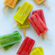 Colorful red, yellow, green, and orange popsicles made with the Nama Popsicle Mold are arranged on a white surface, with their wooden sticks visible.