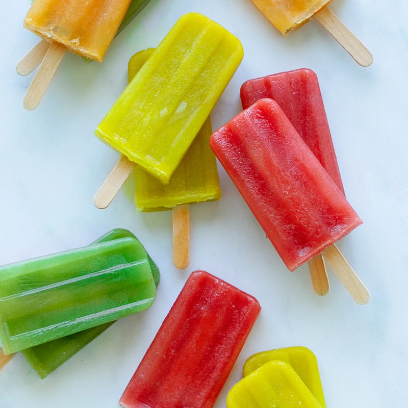 Colorful red, yellow, green, and orange popsicles made with the Nama Popsicle Mold are arranged on a white surface, with their wooden sticks visible.