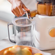 A hand uses the Pulp Strainer to pour no pulp juice into a clear pitcher, while a bowl of oranges rests on the kitchen counter.