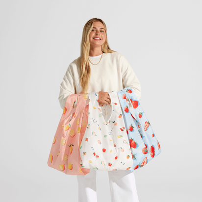 A smiling woman in a cream sweater and white pants holds three Nama + Baggu Standard Tote Bags with fruit patterns, standing against a plain light background.