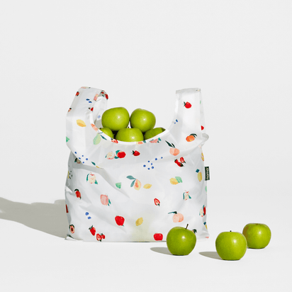 The Nama + Baggu Standard Tote Bag, featuring a colorful fruit design, holds several green apples, with three more green apples placed next to it on a white surface.