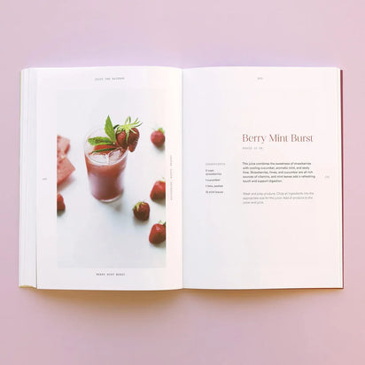 An open recipe book on a pink surface shows a vibrant photo of a pink smoothie garnished with mint and strawberries, surrounded by fresh berries—its “Berry Mint Burst” recipe features Juice the Rainbow for plant-based nutrition.