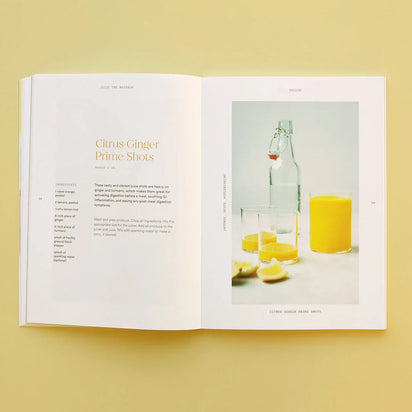 An open recipe book on a yellow surface shows a Juice the Rainbow recipe on one page and, on the other, a photo of vibrant yellow juice with lemon slices and a glass bottle—perfect inspiration for plant-based nutrition enthusiasts.