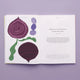 An open Juice the Rainbow recipe book displays a red onion, purple circles, and green leaves on the left page. The right page features info about purple produce, a quote, and a list of plant-based nutrition foods at the bottom.
