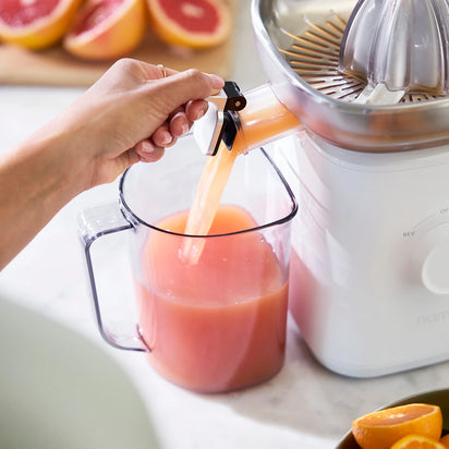 A hand lifts the Citrus Attachment spout, pouring fresh citrus juice into a clear pitcher on a kitchen counter, with halved grapefruits in the background.