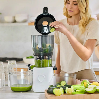 A woman places a cucumber slice into the Nama J2 Juicer’s Large Hopper in her kitchen, with cut vegetables and a glass pitcher of green juice nearby—perfect for easy batch juicing.