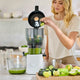 A woman places a cucumber slice into the Nama J2 Juicer’s Large Hopper in her kitchen, with cut vegetables and a glass pitcher of green juice nearby—perfect for easy batch juicing.