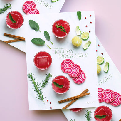 A cookbook titled A Collection of Holiday Recipes rests on a pink surface, surrounded by festive ingredients—red drinks, rosemary, herbs, beet slices, lime, cinnamon sticks, and pomegranate seeds—for healthy holiday treats and plant-based drinks.